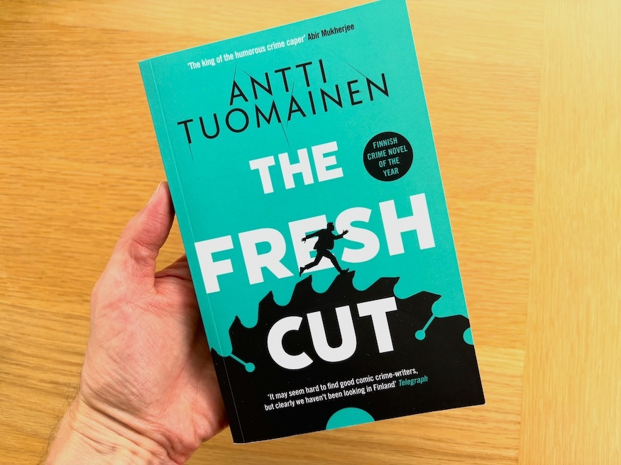 Hand holding The Fresh Cut, a novel by Antti Tuomainen, from Finland